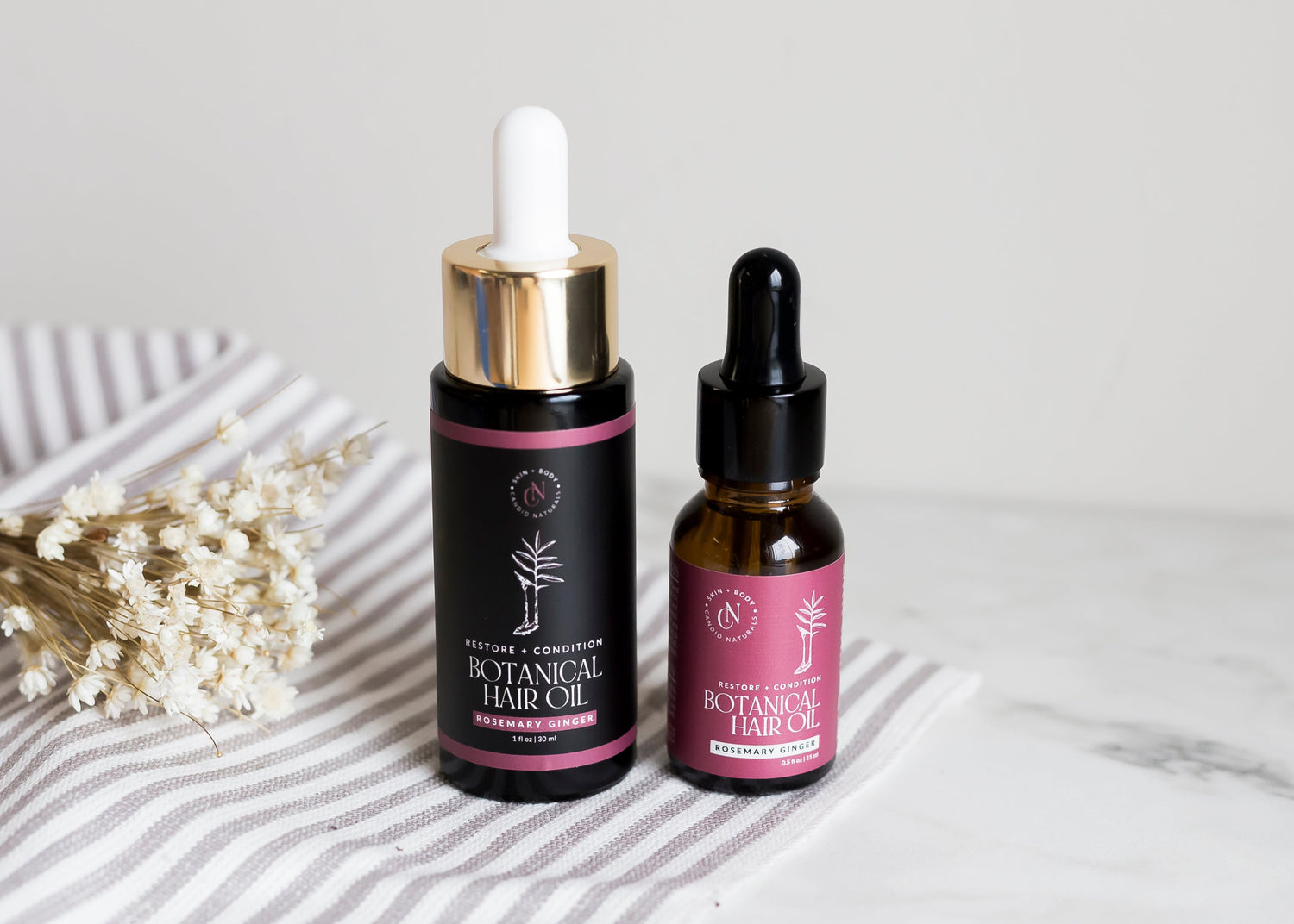 Two bottles of botanical hair oil with droppers on a marble surface.