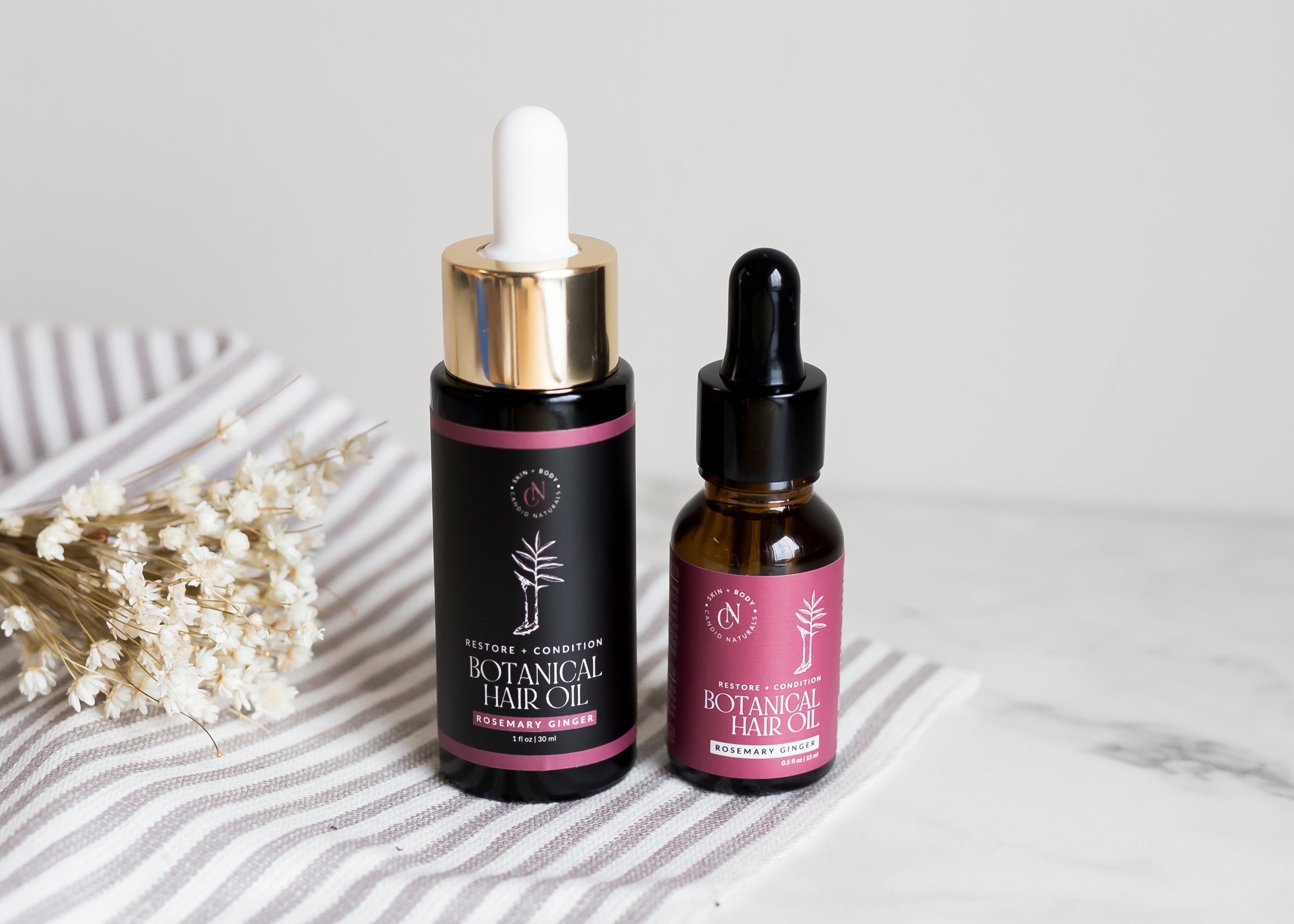 Two bottles of botanical hair oil with droppers on a marble surface.